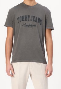 Šedé bavlněné tričko s kulatým výstřihem a krátkým rukávem. Obsahuje velký tištěný logo "TOMMY JEANS New York" v tmavě šedém písmu.