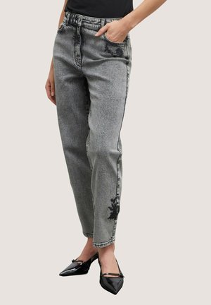 Kobieta nosząca szare dżinsy z wysokim stanem w stylu acid wash z czarnymi kwiatowymi plamami, czarny top i czarne lakierowane buty na obcasie z szpiczastymi noskami.