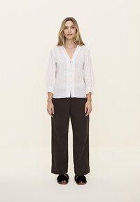 Blouse en lin blanc avec un décolleté en V et une fermeture à boutons, dotée de manches bouffantes avec des poignets froncés. Le modèle l'a associée à un pantalon large sombre.