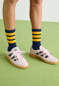 Ανοιχτό ροζ σουέντ αθλητικά παπούτσια με μαύρες ρίγες και καουτσούκ σόλες, συνδυασμένα με ναυτικές και κίτρινες ριγέ ριμπ κάλτσες. Πράσινο υφασμάτινο δάπεδο.