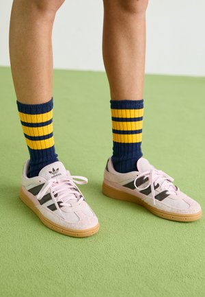Jasnoróżowe zamszowe sneakersy z czarnymi paskami i gumowymi podeszwami, zestawione z niebiesko-żółtymi skarpetkami w paski o ribbowanej strukturze. Zielona fakturowana podłoga.
