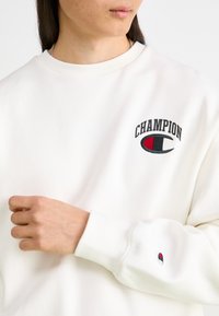 Bijela dukserica s okruglim izrezom, ukrašena izvezenim tekstom "CHAMPION" i logotipom u crnoj i crvenoj boji na prsima i rukavu.