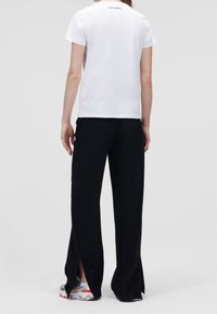 Camiseta blanca de algodón y pantalones negros texturizados con cortes laterales, combinados con zapatillas multicolores. Diseño minimalista, sin logotipos visibles en la parte delantera.