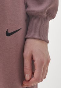 Långärmad mauve sweatshirt med ribbade muddars och en svart broderad Nike-logotyp på framsidan, med en hand som vilar mot tyget.
