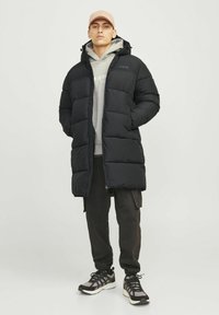 Abrigo de plumas negro con cierre frontal y capucha, que presenta un diseño acolchado. Combinado con una sudadera gris y pantalones oscuros, complementado con zapatillas.
