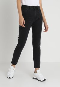 Jeans de mezclilla negros con corte ajustado, diseño de cinco bolsillos y detalles de costura sutiles. Combinados con zapatillas blancas para un contraste.