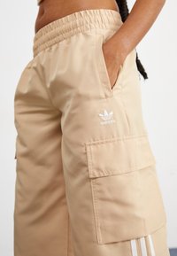 Μπεζ cargo παντελόνια με ελαστική μέση, πλαϊνές τσέπες και λευκό λογότυπο Adidas. Διαθέτουν λευκές ρίγες στα πλάγια και μαλακή υφή.