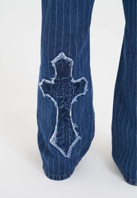 Jeansy z rozkloszowanym dołem w niebieskie paski, z przetarciem w kształcie krzyża na kolanie. Krawędzie są postrzępione dla dodatkowej tekstury.