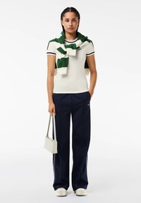 Polo blanco de manga corta con ribete en azul marino, suéter a rayas verde y blanco drapeado sobre los hombros, pantalones anchos en azul marino y bolso claro.