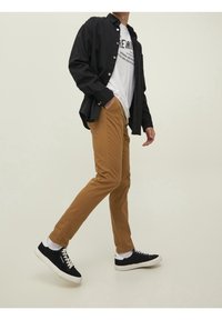 Bruna slim-fit byxor kombinerade med en svart knapp-up skjorta och en vit t-shirt med tryck. Svarta sneakers med vita detaljer. Slät tygtextur.