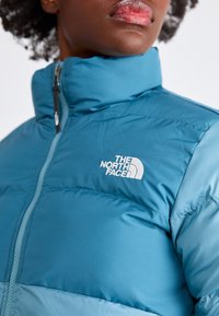 Mėlynas pūkinis džemperis su aukštu apykakle, turintis užtrauktuką ir išsiuvinėtą baltą logotipą krūtinėje. Lygus audinys ir šiltas dizainas.