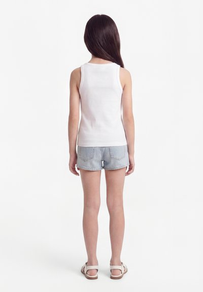 Fille aux longs cheveux bruns, vue de dos, portant un débardeur blanc, un short en jean clair et des sandales blanches.