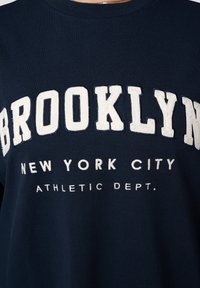 Sweat-shirt bleu marine à col rond avec du texte brodé blanc : "BROOKLYN", "NEW YORK CITY" et "ATHLETIC DEPT." sur le devant.