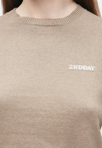 Béžový pletený svetr s kulatým výstřihem, zdobený jemnou texturou a malým bílým logem "2ND DAY" na přední straně.