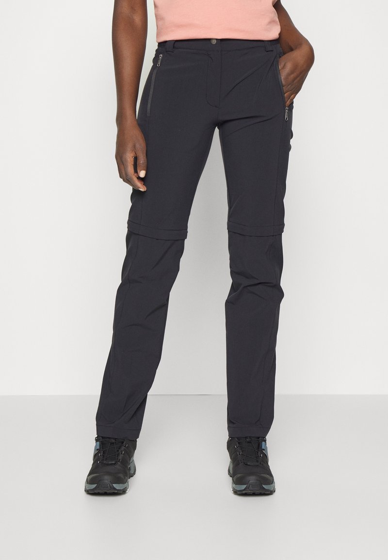 Personne vêtue d’un pantalon de randonnée noir à zip amovible et de chaussures de randonnée noires, debout avec une main dans la poche devant un fond uni.