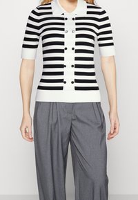 Maglione a righe bianco e nero con maniche corte, colletto e chiusura frontale con bottoni, abbinato a pantaloni grigi sartoriali.