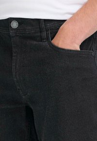 Jeans de mezclilla negra con una vista de cerca, que presentan un bolsillo frontal, una textura sutil y un botón de metal con detalle de marca.