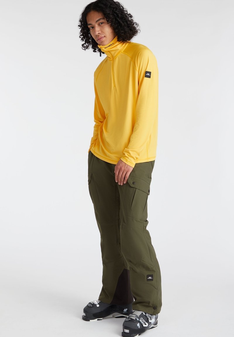 Mann in einem gelben Langarmshirt, olivgrünen Cargo-Hosen und schwarzen Skischuhen, stehend vor einem weißen Hintergrund.