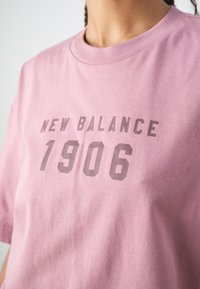 Roza bombažna majica s krogim izrezom in kratkimi rokavi. Na majici je v sivi barvi natisnjen napis "NEW BALANCE 1906", kar kontrastira z barvo majice.