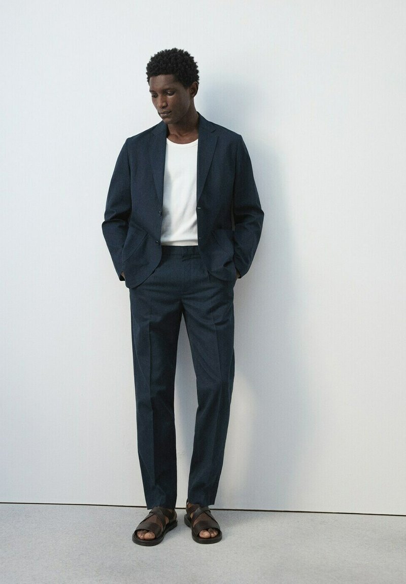 Hombre de pie contra una pared blanca, vistiendo un traje azul marino, camiseta blanca y sandalias marrones, con las manos en los bolsillos y mirando hacia abajo.