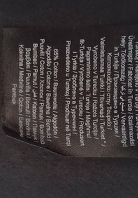 Etiqueta de tela negra con texto blanco. Contiene instrucciones de cuidado y composición del material: 65% algodón, diversas traducciones en varios idiomas.