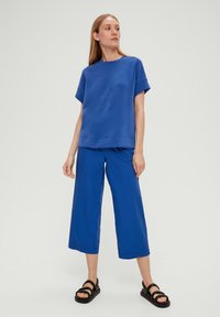 Blauwe katoenen sweatshirt met korte mouwen en reliëfbladontwerp in combinatie met blauwe wijde broeken. Gemaakt met zwarte sandalen.