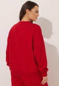 Sudadera roja con corte relajado, escote redondo, puños y dobladillo acanalados. Textura suave, sin patrones visibles ni accesorios.