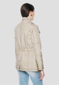 Beige, let jakke med en figursyet talje, struktureret design, synlige sømme og bælte på ryggen. Har ståkrave og sideslids.