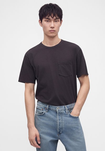 Jeune homme aux cheveux noirs portant un t-shirt noir uni à manches courtes avec poche et un jean bleu clair, une main dans la poche.