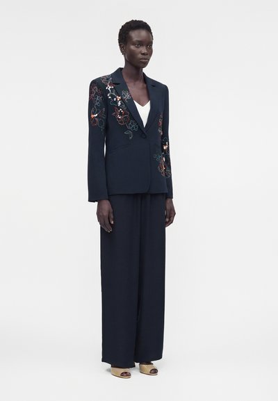 Γυναίκα με ναυτικό σακάκι διακοσμημένο με πολύχρωμη κεντητή λουλουδένια διακόσμηση, φαρδιά ναυτικά παντελόνια, λευκό τοπ από μέσα και μπεζ ανοιχτά δερμάτινα τακούνια, στέκεται ακίνητη.