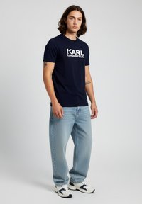 Nuori mies, jolla on pitkät hiukset, päällään laivastonsininen Karl Lagerfeld -T-paita, vaaleansiniset farkut ja valkoiset sekä laivastonsiniset lenkkarit, seisomassa yksinkertaista taustaa vasten.