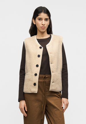 Μπεζ γιλέκο από φλις με στρογγυλή λαιμόκοψη, έξι μπλε κουμπιά και δύο μπροστινές τσέπες, φορεμένο πάνω από καφέ μακρυμάνικο πουκάμισο και καφέ παντελόνι.