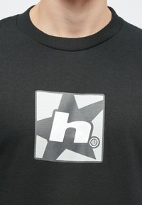 Μαύρο φούτερ με μεγάλο γραφικό σε γκρίζο χρώμα που απεικονίζει ένα στυλιζαρισμένο γράμμα "h" και σχέδιο αστεριού, στρογγυλή λαιμόκοψη, και ραμμένα μανίκια.