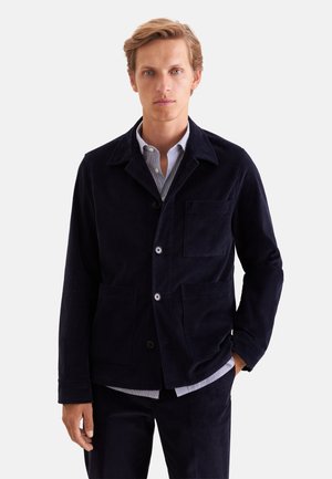 Un homme portant une veste en velours côtelé bleu marine foncé et un pantalon assorti par-dessus une chemise à rayures se tient devant un fond blanc uni.