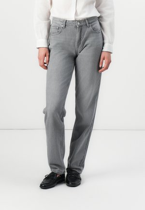 Een persoon staat tegen een neutrale achtergrond, gekleed in een witte blouse en grijze jeans, gecombineerd met zwarte schoenen. Hun handen hangen ontspannen naast hun lichaam.