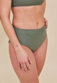 Kobieta w oliwkowych, wysokich majtkach bikini oraz topie, stojąca z ręką opartą na biodrze, z małymi tatuażami na palcach i nadgarstku.