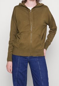 Olivově zelená mikina na zip s kapucí na stažení, dvěma předními kapsami, žebrovanými manžetami a lemem. Měkká textura s neformálním designem.