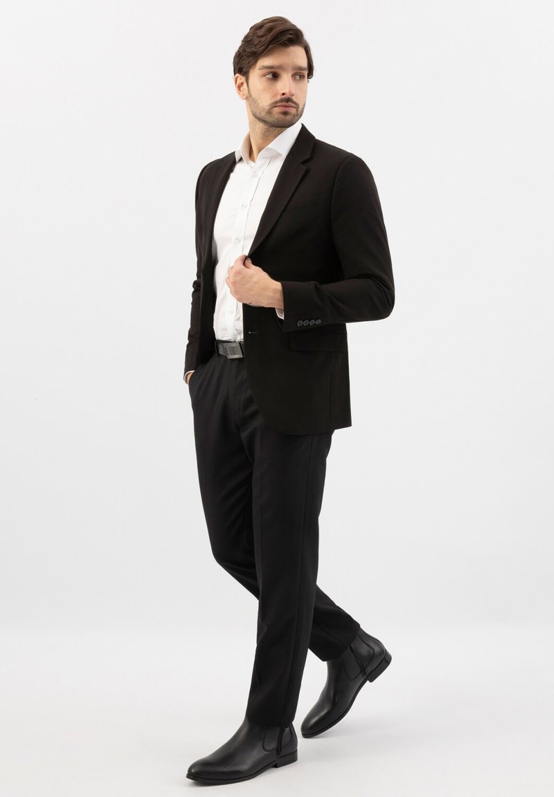 Homme portant un costume noir, une chemise blanche et des bottes noires, debout avec une main dans la poche et l'autre ajustant sa veste.