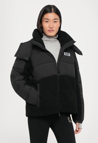 Zwarte pufferjack met een fleece ondersectie, ritssluiting, grote kraag en logo-embleem. Heeft een boxy pasvorm en knoopdetails op de schouders.