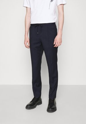 Homme portant un pantalon slim bleu marine avec taille à cordon, associé à des chaussures en cuir noires à lacets et une chemise blanche à manches courtes.