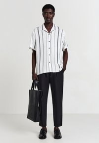 Weißes Kurzarmhemd mit vertikalen schwarzen Streifen, schwarze Jogginghose mit Kordelzug und schwarze Ledertasche. Glattes Material, lässige Passform.