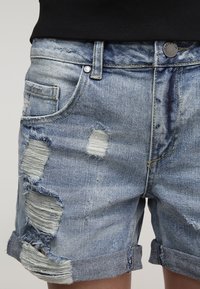 Pantalones cortos de mezclilla azul claro con desgastes, bordes deshilachados y puños doblados. Cuenta con bolsillos tradicionales y cierres de botón metálico.