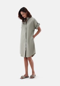 Abito camicia verde chiaro, con bottoni e maniche corte con risvolto. Presenta un orlo arrotondato e tasche laterali. Abbinato a sandali a scivolo coordinati.