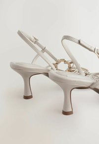Weiße Sandalen mit hohen Absätzen, dünnen Riemen, dekorativen goldenen Akzenten und einem geschwungenen Absatzdesign. Glatte Ledertextur und weiche Form.