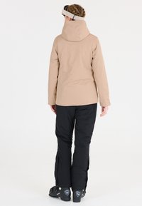 Beige vattentät skidjacka med huva, kombinerad med svarta isolerade skidbyxor och bergsklättringsskor, sedd bakifrån.