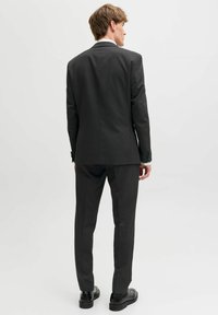 Noor mees seisab seljaga vaataja poole, kandes musta ülikonda koos sobivate pükste ja mustade kingadega, taustal ühtlane valge pind.