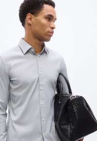 Νέος άνδρας με ανοιχτό γκρι πουκάμισο με κουμπιά που κρατάει μια μαύρη υφαντή δερμάτινη τσάντα, μπροστά σε λευκό φόντο, κοιτάζοντας προς το πλάι.