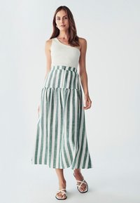 Prúžkovaná maxi sukňa v zelenej a bielej zmesi ľanu, s vysokým pásom a viacvrstvovým dizajnom. Skombinovaná s priliehavým bielym topom na ramienka a sandálmi.