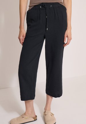 Person iført løse sorte cropped bukser med snøre, taupefarvet skjorte og beige slip-on sko står indendørs på et lyst gulv.