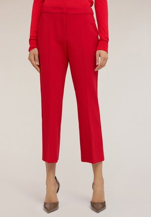 Rote Tailored-Hosen mit glatter Textur, verkürzter Länge und seitlichen Taschen. Dazu werden spitze beige Absatzschuhe für einen Kontrast getragen.